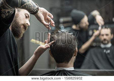 Unmatched barber with a big beard is cutting the hair ends of his client in the black cutting hair cape in the barbershop. Customer has hairgrips on the head. The both blurry reflected in the mirror.