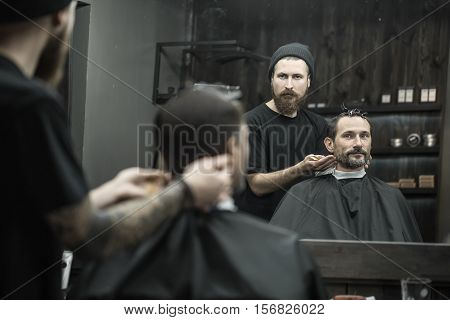 Man and a barber reflected in a mirror in the barbershop. Hairdresser wears a black T-shirt with a cap and has a hair comb. Client wears a black cutting hair cape and has hairgrips on his head.