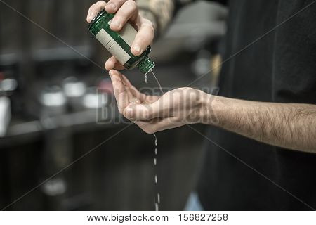 Green bottle with the lotion in the man's hands on the blurry background of the barbershop. Guy is pouring the lotion on his hand. Closeup. Horizontal.