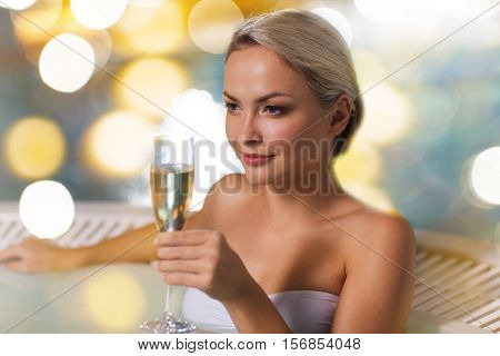 people, beauty, spa, healthy lifestyle and relaxation concept - beautiful young woman wearing bikini swimsuit sitting with glass of champagne in  at poolside over holidays lights background