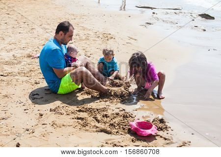 Kids burying dad in sand. UV and sun protection rashguards on the father and children. Games on the beach, sunshine