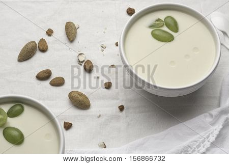 Two white bowls with white garlic soup with three pieces of grapes on a white tablecloth with raw almonds.