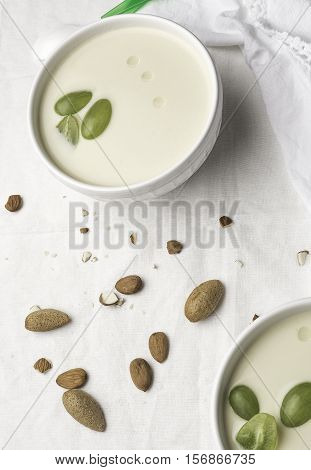 Two white bowls with white garlic soup with three pieces of grapes on a white tablecloth with raw almonds.