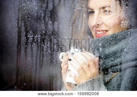 attractive adult woman drinking coffee and looking out of the window on rainy day, close up portre in winter time