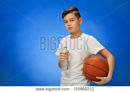 Adorable 11 year old boy child with basketball ball drinking water on blue background.
