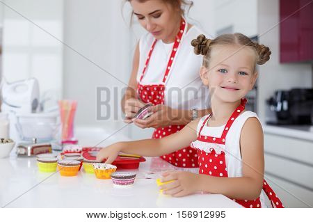 Young woman,brunette,hair gathered on the crown,dressed in a white shirt and a red apron with white polka dots,with my daughter 5 years old,a little blond girl in white tank top and a red apron with white polka dots baking cupcakes in the kitchen
