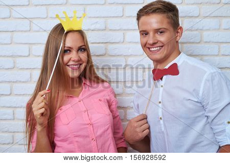 Close-up shot of extremely elated couple while standing over white background.  Funny couple holding paper bow-tie and crown, they are laughing while posing with paper accessories