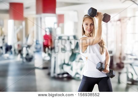 Young beautiful girl makes exercises with weight in gym. Blurred background