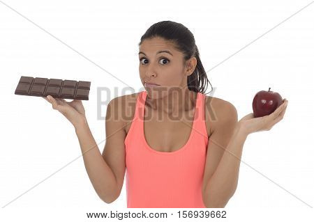 young attractive hispanic woman in fitness top holding apple fruit and chocolate bar in her hands in diet dilemma sweet tasty unhealthy food against healthy nutrition choice