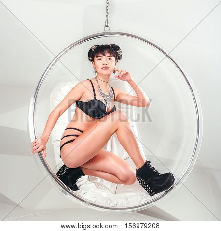 Beautiful slim model woman in underwear posing standing in suspended transparent chair in white studio.