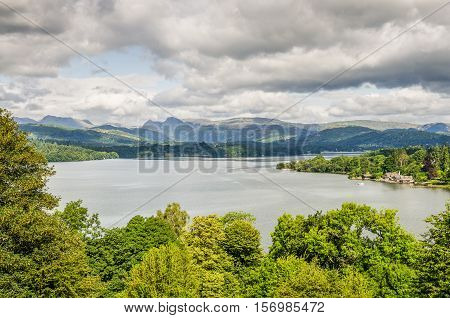 View of Lake Windermere from the south surrounded by trees and distant mountains under a mostly gray sky.
