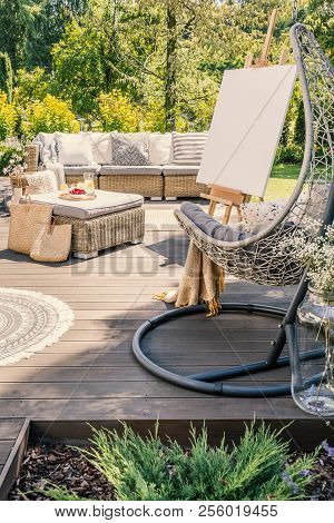 Hanging Chair On Wooden Patio With Easel And Rattan Table In Front Of Sofa In The Garden