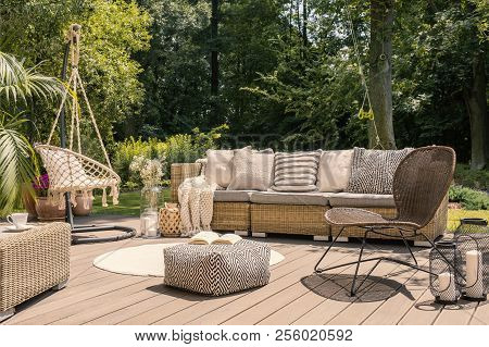 Pouf And Rattan Chair On Wooden Patio With Settee In The Garden During Summer. Real Photo