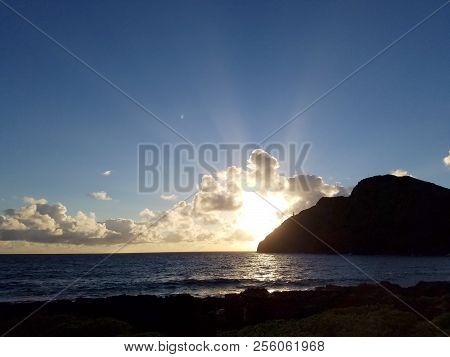 Ocean Waves Lap On Makapuu Beach As Sunrises Through The Clouds With Makapuu Lighthouse And Point In