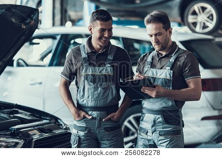 Two Handsome Happy Auto Mecanics In Garage. Portrait Of Attractive Repairmen In Grey Uniform Showing