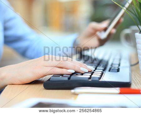 Closeup Female Hands Typing On Keyboard Using Smart Phone At The Desk In The Office.