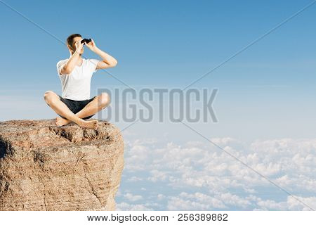 Young Man Using Binoculars On Cliff. Sky Background. Research And Relax Concept