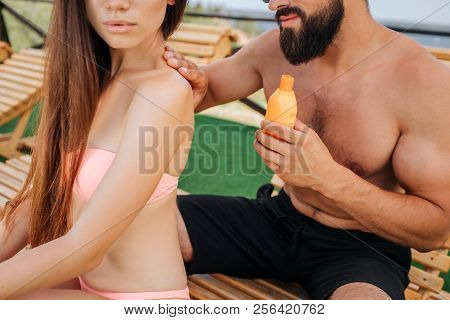 Cut View Of Man And Woman Sitting. She Looks Straight Forward. Guy Puts Some Sunproof Lotion On Her 