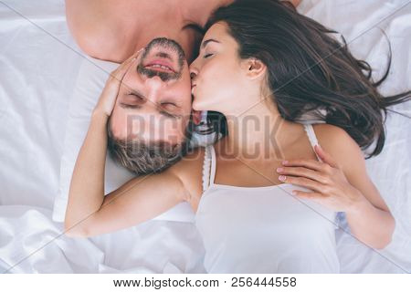 Beautiful Man And Woman Are Lying In White Bed. Guy Keeps Eyes Closed And Smiles. Girl Is Touching H