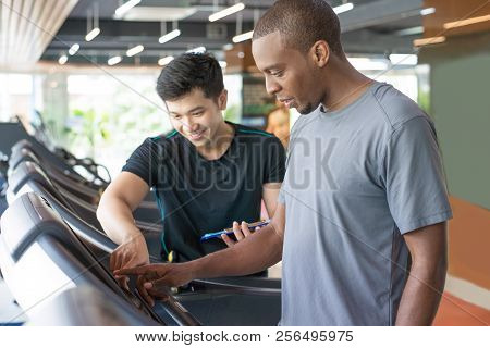 Black Man Setting Up Exercise Machine With Personal Trainer. Serious Young Guy Training In Gym. Work