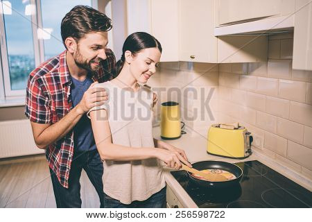 Positive Couple Stands In Kitchen. She Holds Pen Of Pan. Girl Cooks Omlet. Guy Stands Behind Her And