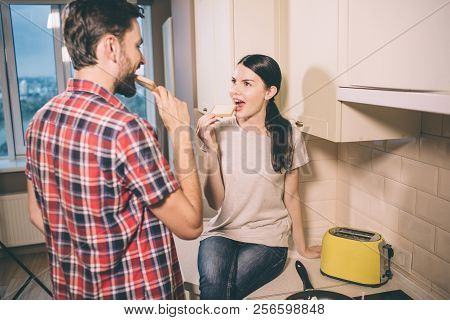 Hungry People Are In Kitchen. Girl Sits And Botes Toast. Guy Looks At Her And Does The Same Thing. T