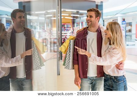 Young Woman With Shopping Bags Pointing By Finger At Shopwindow To Boyfriend At Mall