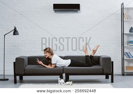 Happy Young Woman Using Smartphone While Lying On Sofa Under Air Conditioner Hanging On Wall