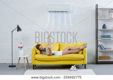 Happy Young Woman Reading Book On Couch And Pointing At Air Conditioner Hanging On Wall With Remote 