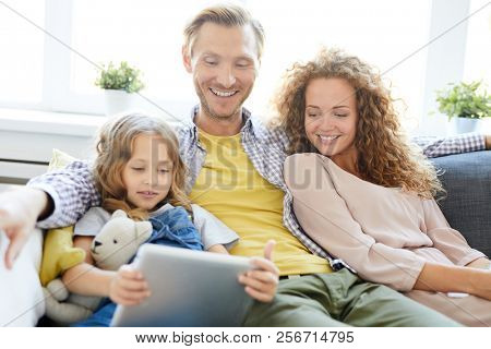 Cute girl with tablet watching curious and funny vids while sitting on sofa by her parents