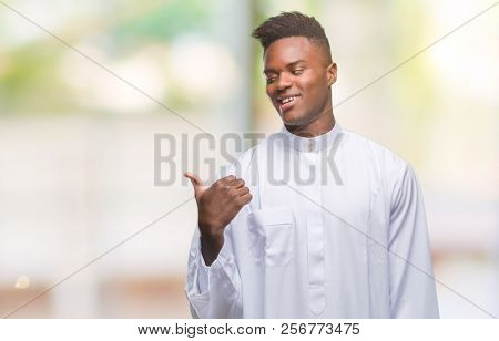 Young arabic african man wearing traditional djellaba over isolated background smiling with happy face looking and pointing to the side with thumb up.