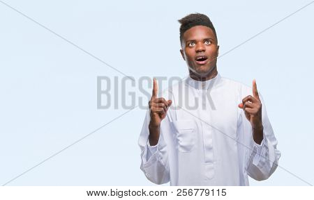 Young arabic african man wearing traditional djellaba over isolated background amazed and surprised looking up and pointing with fingers and raised arms.