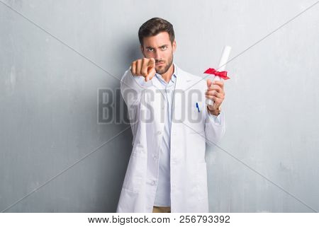 Handsome young doctor man over grey grunge wall holding diploma pointing with finger to the camera and to you, hand sign, positive and confident gesture from the front