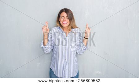 Young adult woman over grey grunge wall wearing fashion business outfit smiling crossing fingers with hope and eyes closed. Luck and superstitious concept.