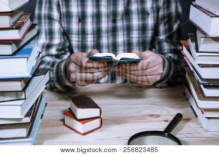 A Man Reading A Miniature Book Among The Big Books In The Library At The Wooden Table.