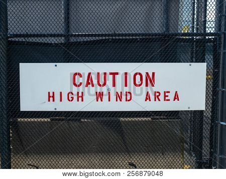 A Caution High Wind Area Sign Handing On Black Fence
