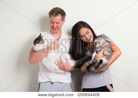 Young Happy Multi-ethnic Couple Holding Two Cats Together Against White Background