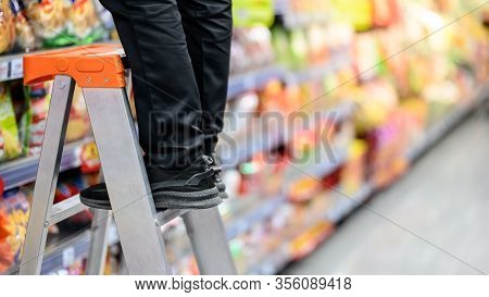 Male Storekeeper Legs Standing On Aluminum Stair Putting Products On Shelves In Grocery Store Or Sup