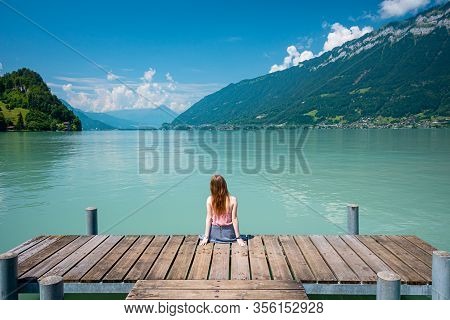 Back View Of A Young Girl Sitting On A Pier. Beautiful Turqouise Lake, Mountains And Sky Background.