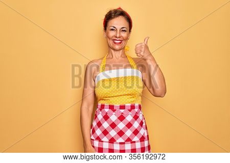 Middle age senior pin up housewife woman wearing 50s style retro dress and apron doing happy thumbs up gesture with hand. Approving expression looking at the camera showing success.