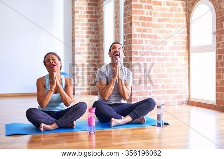 Middle age sporty couple sitting on mat doing stretching yoga exercise at gym begging and praying with hands together with hope expression on face very emotional and worried. Asking for forgiveness