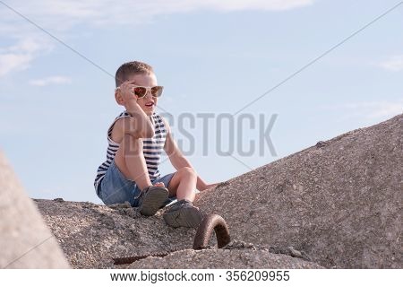 Little Caucasian Boy In Sailor Singlet And Sunglasses Siting On Breakwater Block On The Background O