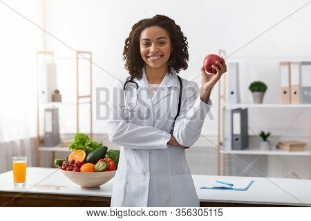 Beautiful African Woman Dietologist Holding Apple In Her Hand, Recommending Fresh Fruits, Posing At 