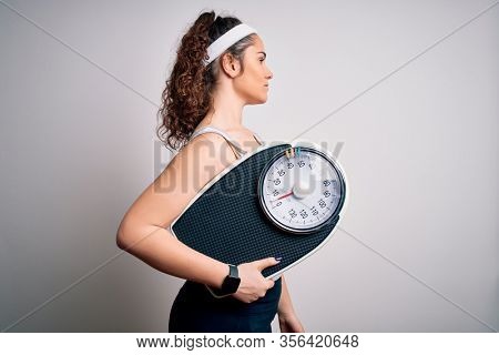 Young beautiful woman with curly hair holding weighing machine over white background looking to side, relax profile pose with natural face and confident smile.