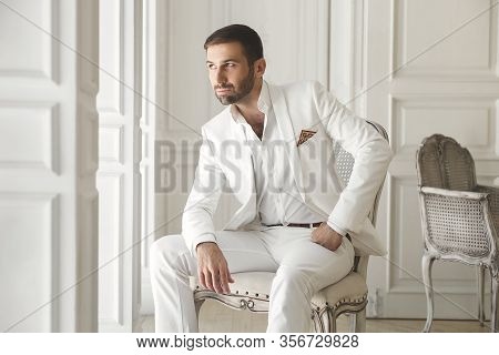 Elegant Young Handsome Man With A Beard In A White Classic Suit. The Hotels Interior. Photo Studio. 