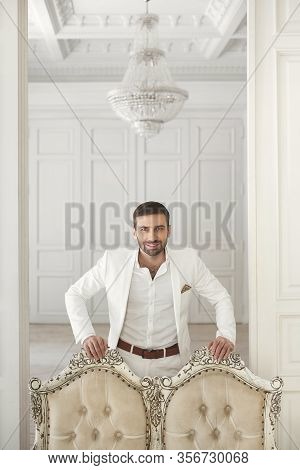 Elegant Young Handsome Man With A Beard In A White Classic Suit. The Hotels Interior. Photo Studio. 