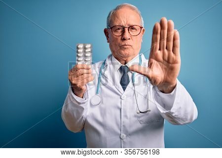 Senior grey haired doctor man holding pharmaceutical pills over blue background with open hand doing stop sign with serious and confident expression, defense gesture