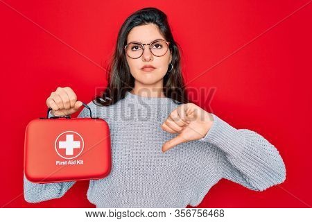 Young beautiful girl holding first aid kit medical box over red background with angry face, negative sign showing dislike with thumbs down, rejection concept