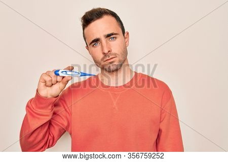 Young handsome man with blue eyes measuring temperature using digital thermometer thinking attitude and sober expression looking self confident
