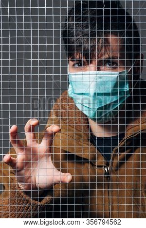 Young Girl Stands Behind A Fence With Medical Mask. It Is In Quarantine. Health And Medicine Concept